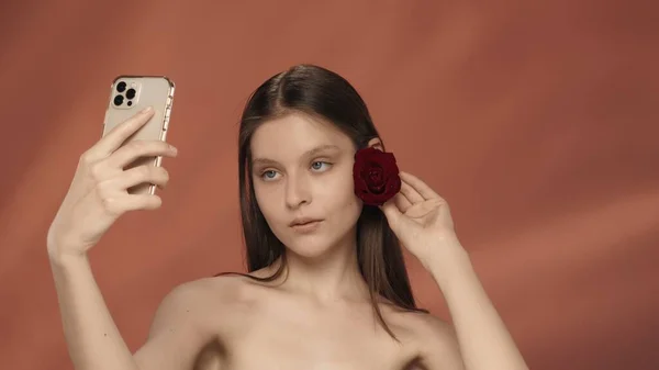 头发上长着红玫瑰芽的女人拍了个视频用智能手机拍了个自拍带着红色背景的女人站在演播室里 美容美发 美容美发 治疗和护理的概念 — 图库照片