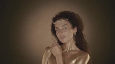 Genç ve çekici bir kadının kameraya bakıp yüzüne dokunduğu videoyu kapat. Altın makyaj, küpeler ve dikenli taç. Altın boya. Kahverengi arka plan. Güzellik ve kozmetik reklamı.