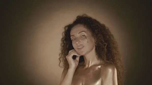 一个年轻迷人的女人看着相机 用手指 金色的化妆品和装饰品触摸着她的脸 金色的水滴棕色背景 化妆品及化妆品广告 — 图库照片