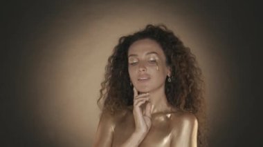 Genç ve çekici bir kadının kameraya baktığı ve yüzüne parmaklarıyla, altın makyajıyla ve aksesuarlarıyla dokunduğu videoyu kapat. Altın damlalar. Kahverengi arka plan. Güzellik ve kozmetik reklamı