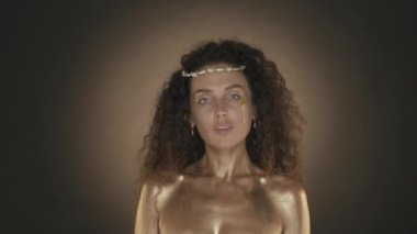 Genç ve çekici bir kadının kameraya bakışının videosunu çek, kalçasında altın bir gözyaşı var. Altın makyaj ve dikenli taç. Altın boya. Kahverengi arka plan. Güzellik ve kozmetik reklamı. HDR