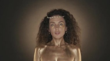 Genç ve çekici bir kadının kameraya baktığı, yanaklarında altın gözyaşları olan videoyu kapat. Altın makyaj ve dikenli taç. Altın boya. Kahverengi arka plan. Güzellik ve kozmetik reklamı