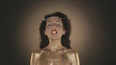 Genç ve çekici bir kadının kameraya bakıp şeker yediği videoyu kapat. Altın makyaj, küpeler ve dikenli taç. Altın boya. Kahverengi arka plan. Güzellik ve kozmetik reklamı. HDR