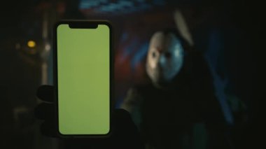 Hokey maskeli bir adam elektrikli testere tutuyor ve kameraya akıllı telefonu gösteriyor, depoda duruyor. Bir erkek modelin portresi. Ekrana odaklan. Reklam alanı, çalışma alanı modeli.
