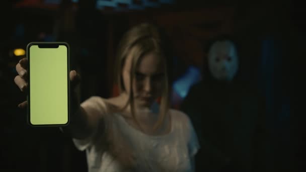 恐怖狂人的女受害者 站在那里拿着智能手机 在摄像机前竖起大拇指 一个戴着冰球面具的男人站在她身后的旧仓库里 广告区 工作空间模型 — 图库视频影像