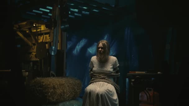 ホラー映画シーン 倉庫の椅子に座っている白いドレスのマニアックのブロンドの女性の犠牲者 彼女はロープで縛られている ホッケーマスクの男はチェーンソーで彼女の前に立っています — ストック動画