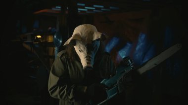 Korku filmi sahnesi. Domuz maskesi takan ve elinde testere tutan bir adam eski deponun içinde duruyor. Çirkin maskeli bir erkek modelin yakın portresi. Cadılar Bayramı konsepti.