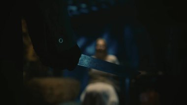 Korku filmi sahnesi. Yakın çekim, bıçağa odaklan. Arka planda beyaz elbiseli bir manyağın sarışın kadın kurbanı depoda bir sandalyede, iple bağlanmış olarak oturuyor..