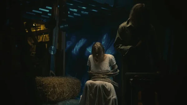 恐怖的电影场景 恐怖狂热者的受害者坐在仓库里的椅子上 用绳子捆着 男人拿着骡子 摸着姑娘 吓着她 — 图库照片