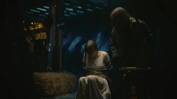ホラー映画シーン 白い長いドレスの女性の犠牲者は ロープで縛られた倉庫の椅子に座っています 彼女の隣に立って話す男のホラーマニアック — ストック写真