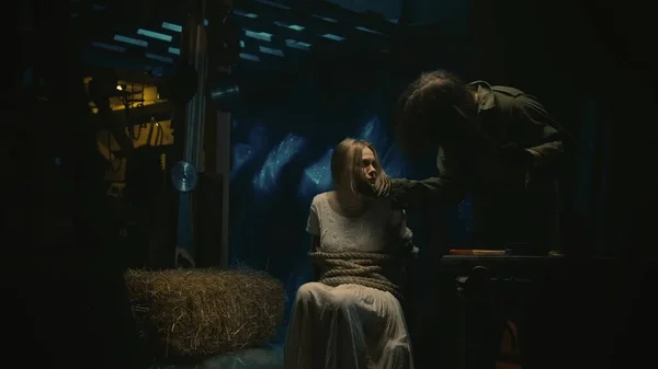 ホラー映画シーン 古い倉庫にロープで縛られた椅子に座っている白い長いドレスの女性の犠牲者 彼女の顔に触れる彼女の隣に立っている男のホラーマニアック — ストック写真