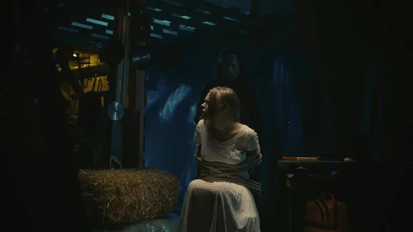 恐怖的电影场景 穿着白衣 坐在仓库椅子上的金发女孩成为了这个疯子的牺牲品 她被绳子捆住了 戴着冰球面具的男人站在她身后 — 图库照片