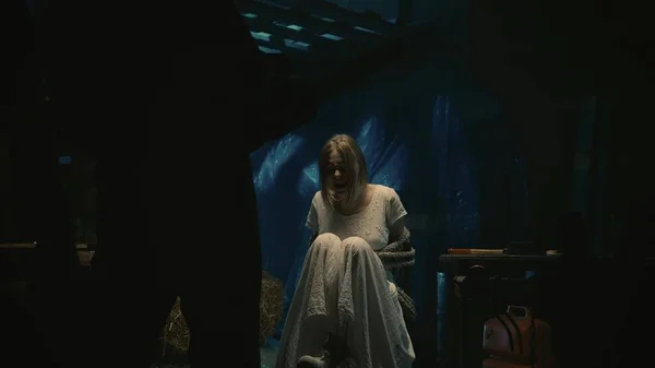 恐怖的电影场景 一个金发女模特儿受害者穿着连衣裙坐在仓库里的椅子上 用绳子捆着 拍了一张特写镜头 前面站着一个拿着刀的男人 — 图库照片