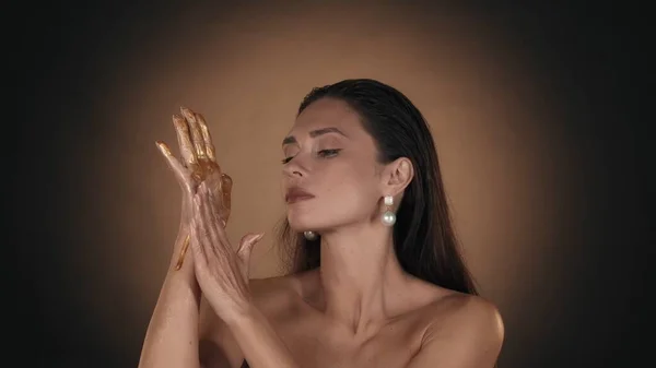 Close up shot of a female model with pearl earrings looking at the golden paint flowing down from her hand and touching it. Beauty advertisement concept.