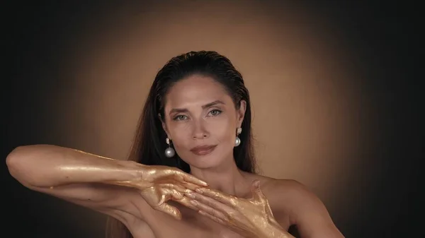 Portrait of a beautiful woman. Close up shot of female model with shiny makeup applying golden liquid paint over her hands skin. Beauty advertisement concept.