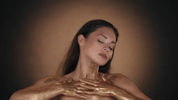 Portrait of a appealing woman with glowing makeup. Close up shot of female model applying golden liquid paint over her chest area. Beauty advertisement concept.