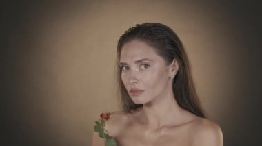 Çekici esmer kadının portresi. Stüdyoda, elinde gül çiçeği olan ve kameraya bakarak vücuduna altın serumu uygulayan bir kadın modelin çekimini kapat. Güzellik reklamı konsepti. HDR BT2020