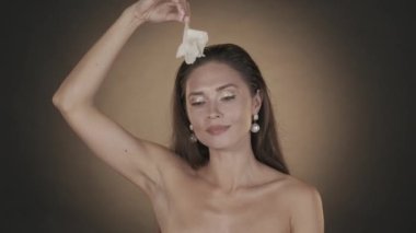 Çekici esmer kadının portresi. İnci küpeli, beyaz çiçekle oynayan ve kameraya bakan kadın modelin stüdyo fotoğraflarını çek. Mücevher reklamı konsepti. Yavaş çekim HDR