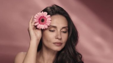Bir kadın kusursuz cildinde Gerbera çiçeği taşıyor. Stüdyoda pembe arka planda Gerbera çiçeği olan bir kadının portresi. Kozmetik ürünleri, çiçek özlü ve güzel kokulu. Şey...