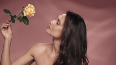 Bir kadın boynundaki gül çiçeğini çıplak omuzlarından geçirir kozmetik etkisinden zevk alır. Stüdyoda pembe arka planda bir kadın var. Güzellik konsepti, kozmetoloji, cilt bakımı