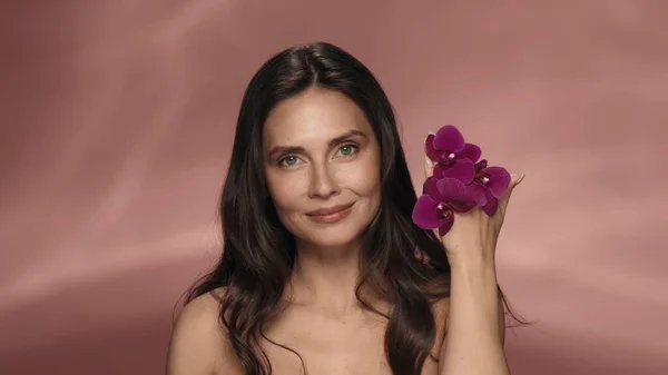 彼女の手に紫色の蘭の花が付いている女性の肖像画が閉じます ピンクの背景にあるスタジオのセミノードの女性 オーキッドエキスで手スキンケアのための化粧品ライン 美のコンセプト — ストック写真