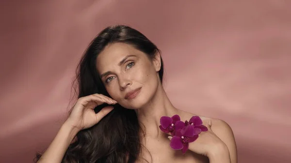 ピンクの背景にあるスタジオで紫色の蘭の花を手にしたセミナーの女性の肖像画 女性は顔と体の皮膚に触れ そのベルベットを楽しんでいます 美のコンセプト — ストック写真