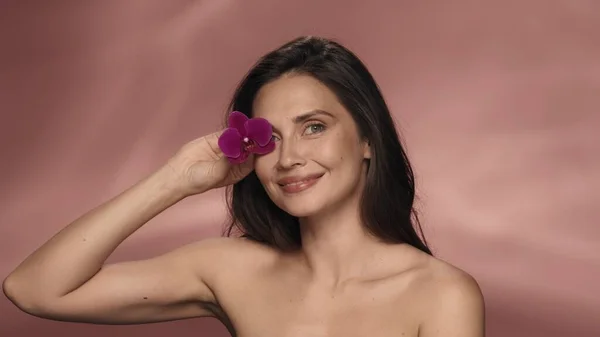 セミナーの魅力的な女性は紫色の蘭の花で彼女の目をカバーしています ピンクの背景にあるスタジオにいる女性 オーキッドエキスと顔の皮膚と目の輪郭のための化粧品ライン — ストック写真