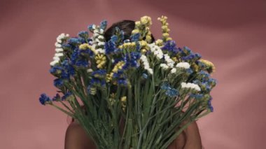 Bir kadın yüzünü bir buket kır çiçeğinin arkasına saklar. Kadın buketi itiyor ve mutlu bir şekilde gülümsüyor. Stüdyoda pembe arka planda genç bir kadın var. Güzellik kavramı