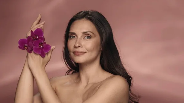 一个手上拿着紫色兰花的女人的画像 粉红背景的演播室里显赫的女人 用兰花提取物护理手部皮肤的化妆品线 美的概念 — 图库照片