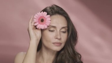 Bir kadın kusursuz cildinde Gerbera çiçeği taşıyor. Stüdyoda pembe arka planda Gerbera çiçeği olan bir kadının portresi. Kozmetik ürünleri, çiçek özlü ve güzel kokulu. Şey...