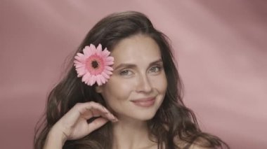 Bir kadın dokunarak ipeksi saçlarından zevk alır. Stüdyoda pembe arka planda saçında Gerbera çiçeği olan bir kadının portresi. Kozmetik saç bakım ürünleri.