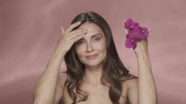 Kadın yüzünü kaplayan orkide çiçekleriyle ellerini çeker ve açar. Bir kadın elini pembe arka planda gezdirirken yumuşak deriden hoşlanır. Orkide özlü kozmetik ürünleri. Şey...