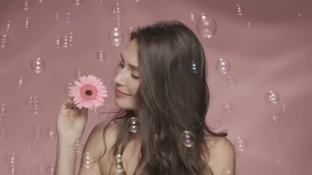 一个无忧无虑的女人 开着一朵花 周围都是肥皂泡 一个女人在工作室的头发上插了一朵粉红色背景的花 女性气质 — 图库视频影像