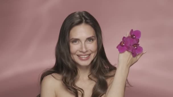 女性は彼女の顔から蘭の花で手を取り除き きれいで滑らかな肌を示します ピンクのバックグラウンドでスタジオのセミナーの女性を魅了 オーキッドエキスを使った化粧品 コンセプト — ストック動画