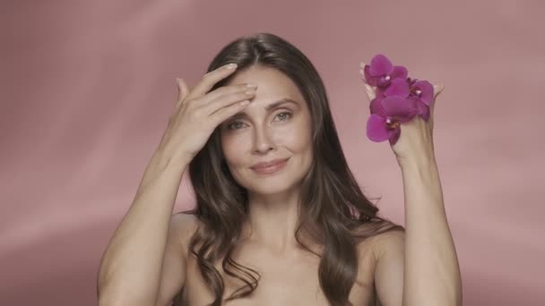女性は彼女の顔を覆う蘭の花で彼女の手を取り除き それを開きます 女性はピンクの背景に彼女の顔の上に手を走らせながら柔らかい肌を楽しんでいます オーキッドエキスを使った化粧品 イギリス — ストック動画