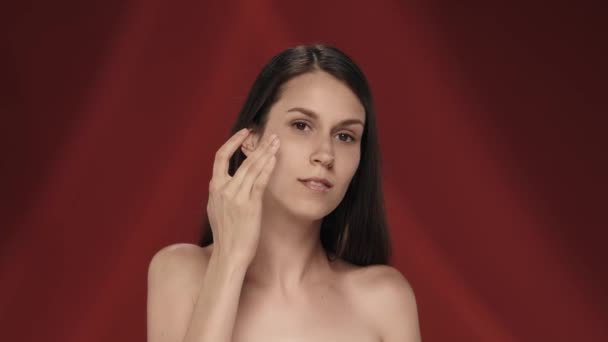여자는 피부를 돌보고 얼굴에 크림을 바릅니다 빨간색 배경에 스튜디오에서 세미나 — 비디오