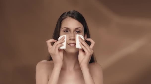 女性は顔を浄化するために綿パッドを使用します 茶色の背景にあるスタジオの若いセミナーの女性の肖像画 ケアのコンセプト — ストック動画