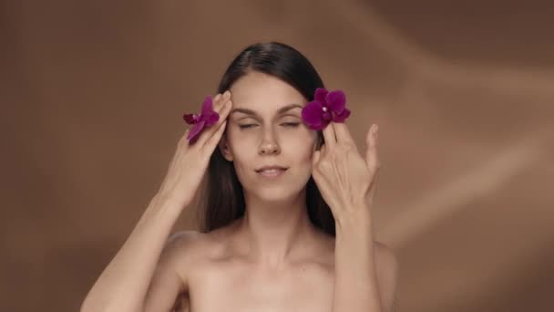 手のひらに蘭の花が咲いている女性が顔に触れ 滑らかで柔らかい肌を楽しんでいます ブラウンの背景にあるスタジオのセミナーの女性の肖像画 美のコンセプト 化粧品 — ストック動画