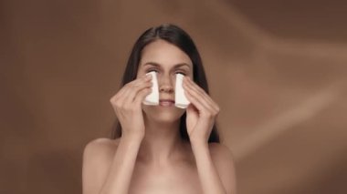 Bir kadın pamuk pedlerin yardımıyla yüzüne krem sürer. Stüdyoda kahverengi arka planda genç bir seminer kadınının portresi. Güzellik, kozmetoloji, özen kavramı. Yavaş çekim