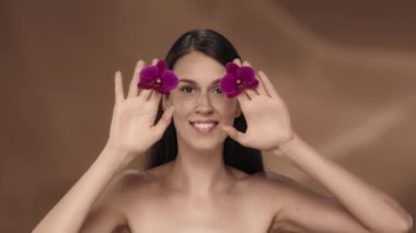Bir kadın ellerini mor orkide çiçekleriyle kapatır, ellerini şaşırmış bir ifadeyle çeker. Stüdyoda kahverengi arka planda bir kadın var. Ürününüzü sunuyorum.
