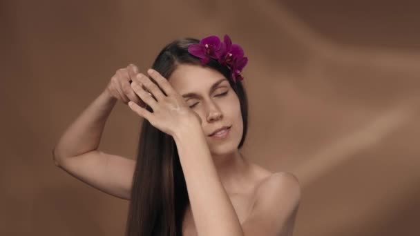 長い髪をかぶっている女性 スタジオで茶色の背景に彼女の髪に蘭の花を持つセミノードの女性 化粧品 ケアのコンセプト スローモーション — ストック動画