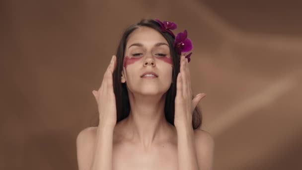 目の下にヒドロゲルパッチが付いている女性は 彼女の顔に触れ ウィンクします 茶色の背景にスタジオで彼女の髪に蘭の花を持つセミノードの女性 美しさ ケアについて スローモーション — ストック動画