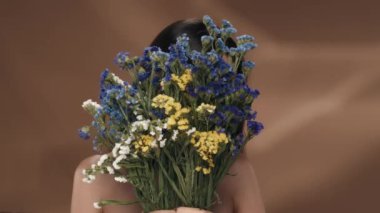 Bir kadın yüzünü bir buket kır çiçeğinin arkasına saklar. Kadın buketi itiyor ve göz kırpıyor. Stüdyoda kahverengi arka planda bir kadın var. Yakın plan. Güzellik konsepti, kozmetoloji