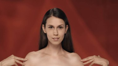 Genç kadın pürüzsüz, ipeksi teninin tadını çıkarıyor, omuzlarına dokunuyor. Stüdyoda kırmızı arka planda bir seminer kadınının portresi. Güzellik konsepti, kozmetoloji, cilt bakımı