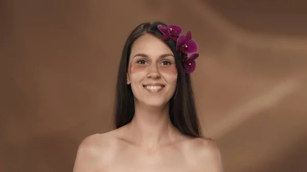 彼女の髪に蘭の花と彼女の目の下のピンクのハイドロゲルパッチが付いているセミナーの女性の肖像画 スタジオの茶色の背景にある女性 美のコンセプト 化粧品 — ストック写真