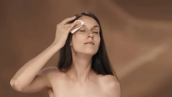 女性は顔を浄化するために綿パッドを使用します 茶色の背景にあるスタジオの若いセミナーの女性の肖像画 ケアのコンセプト — ストック写真