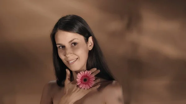 女性がピンクの花で彼女の手に触れ 首の皮膚に触れる 若いセミナーの女性は 茶色の背景にあるスタジオで彼女の完璧な肌を示しています 美のコンセプト 化粧品 — ストック写真