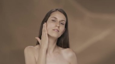 Genç kadın pürüzsüz, ipeksi teninin tadını çıkarıyor, boynuna, omuzlarına ve göğsüne dokunuyor. Stüdyoda kahverengi arka planda gülümseyen bir kadının portresi. Güzellik konsepti, kozmetoloji