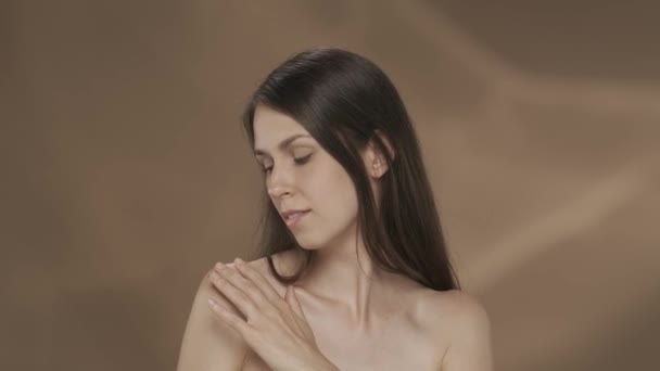 若い女性は彼女の滑らかで きれいな肌を楽しんで 肩や胸に触れています 茶色の背景にあるスタジオのセミナーの女性の肖像画 スキンケアのコンセプト — ストック動画