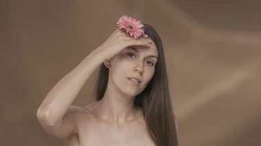 Bir kadın yüzüne, boynuna ve omuzlarına pembe bir gerbera çiçeğiyle dokunuyor. Genç bir seminer kadını mükemmel tenini kahverengi arka planda bir stüdyoda gösteriyor. Konsept: